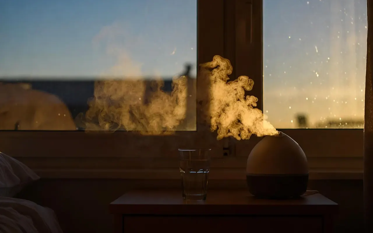 A dim, warm bedroom in the evening, a bedside lamp glowing amber over an open paper book and a mug, the window deep blue with dusk.