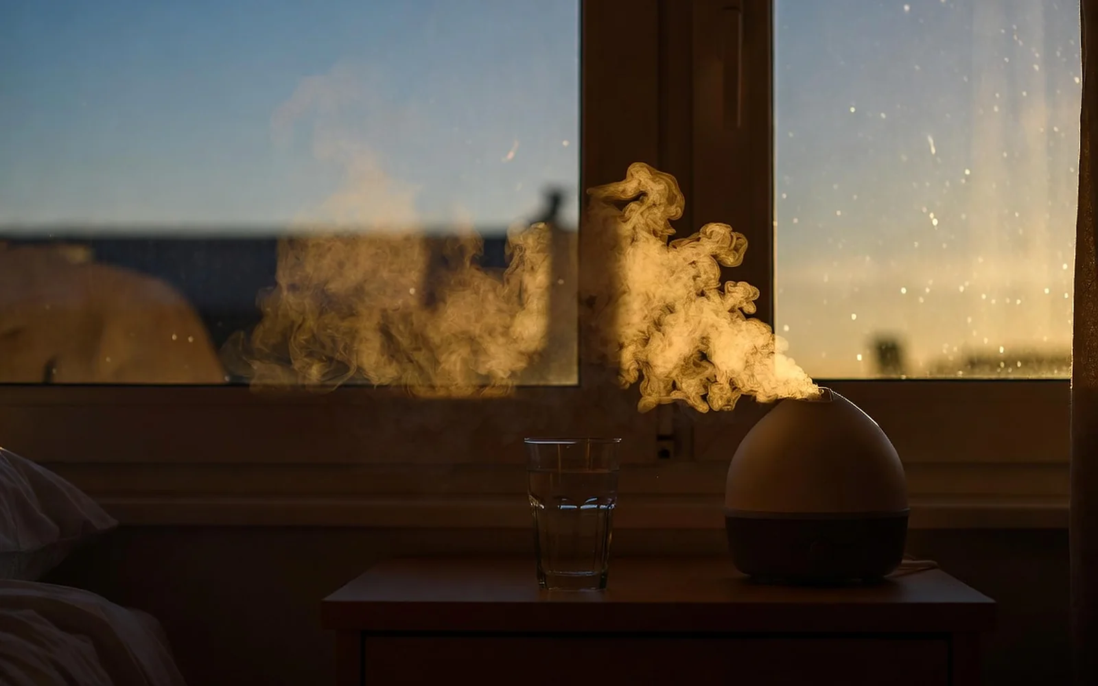 A humidifier on a bedside table sending fine mist into warm evening light, beside a glass of water at the window.
