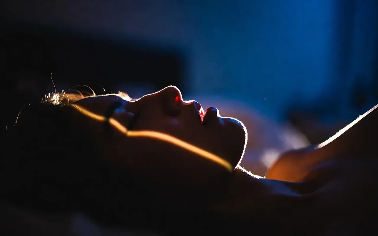 Profile of a person asleep on their side in a dark blue bedroom, face relaxed, edged in warm light.